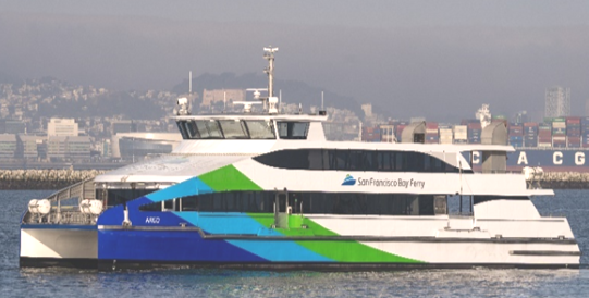 The MV Argo, a ferryboat owned by San Francisco Bay Ferry, sails through the San Francisco Bay. It has a blue, green and white paint job and the San Francisco Bay Ferry logo adorns the side of the vessel. In the background is a large container ship stacked with containers and hills with houses and buildings built onto them.
