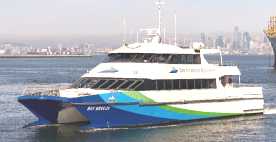 The MV Bay Breeze ferryboat floats atop the water. It has two passenger decks and a blue, green and white paint job. The San Francisco Bay Ferry logo adorns the side. Off in the distance is the San Francisco city skyline.