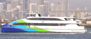 A picture of the MV Carina, a ferry boat in the San Francisco Bay Ferry fleet, sailing across the San Francisco Bay. It has a blue, green and white paint job and the San Francisco Bay Ferry logo adorns the side of the vessel. Distant in the background is the Bay Bridge in front of the San Francisco skyline.