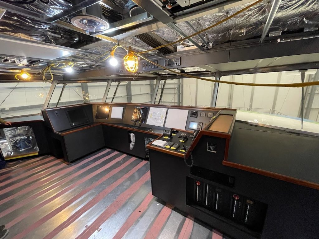 A picture of the control room where the ferry captain will pilot the MV Dorado. There are multiple monitors and control panels. The ceiling is exposed and temporary construction lights hang down providing light for the workers.