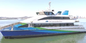 The MV Gemini ferryboat floats atop the water. It has a blue, green and white paint job and the San Francisco Bay Ferry logo adorns the side. The city of San Francisco is distant in the background.