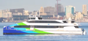 A picture of the MV Hydrus, a ferry boat in the San Francisco Bay Ferry fleet, sailing across the San Francisco Bay. It has a blue, green and white paint job and the San Francisco Bay Ferry logo adorns the side of the vessel. Distant in the background is the San Francisco skyline and Oracle Park, the baseball stadium for the San Francisco Giants.