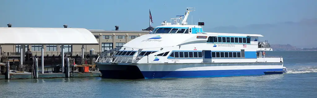 A blue and white ferry boat named M/V Intintoli docks at the San Francisco Ferry Building in downtown San Francisco. It has three logos for the San Francisco Bay Ferry on its hull.