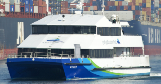 The MV Peralta ferryboat floats atop the water. It has two passenger decks and a blue, green and white paint job. The San Francisco Bay Ferry logo adorns the front and side of the boat. In the background is a large container ship stacked with a variety of containers.