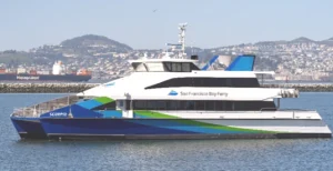 The MV Scorpio ferryboat floats atop the water. It has a blue, green and white paint job and the San Francisco Bay Ferry logo adorns the side. A large container ship is sailing by distant in the background.