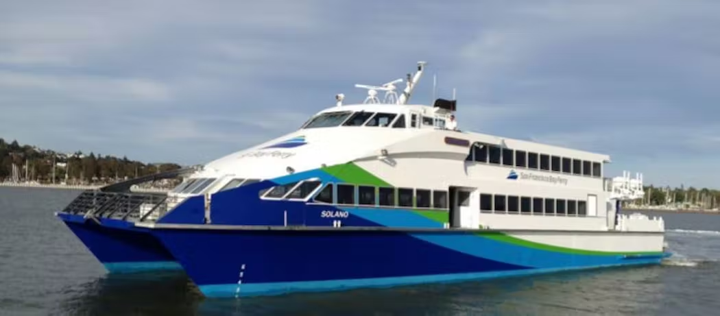A side view of the M/V Solano sailing across the water. The M/V Solano has a blue, green and white paint scheme and the San Francisco Bay Ferry logo adorns its side.
