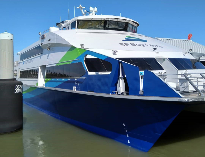 A close up view of the MV Vela, a ferryboat owned by San Francisco Bay Ferry. It has a blue, green and white paint job. The words "SF Bay Ferry" and the San Francisco Bay Ferry logo are on the front of the vessel.