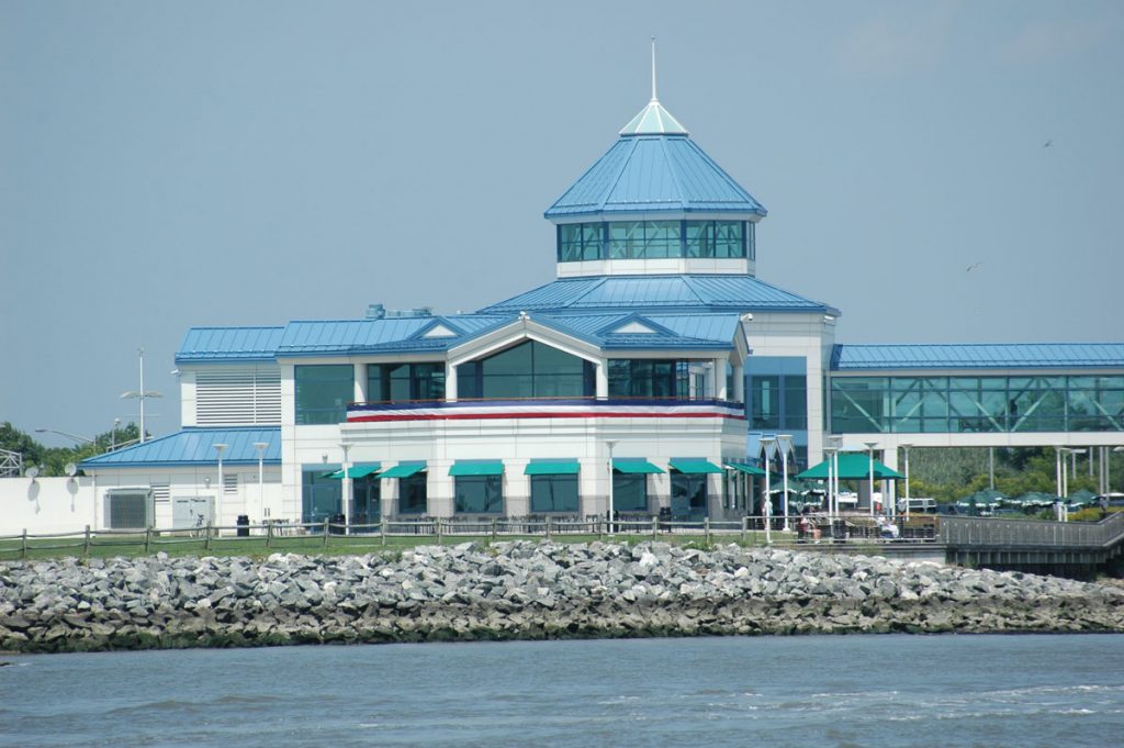 A photo of the Cape May Ferry Terminal taken from a boat in the water. The terminal is two stories and has a light blue metal roof. Green awnings sit atop each window. A pedestrian bridge with floor to ceiling glass is attached to the right side of the terminal.