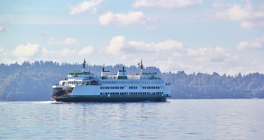 A side view of the MV Issaquah, a auto and passenger ferry operated by Washington State Ferries, departing from Fauntleroy and sailing across Puget Sound. It is painted primarily white with accents of dark green on the hull. In the background is land covered covered in trees in various shades of green.