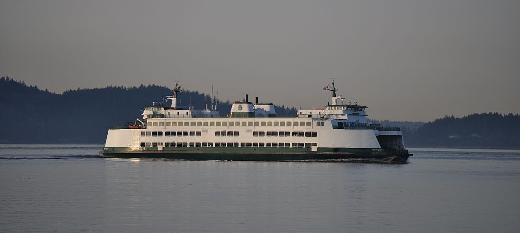 A side view of the MV Issaquah, a auto and passenger ferry owned and operated by Washington State Ferries, sailing across the water north of Vashon Island. It is painted primarily white with accents of dark green along the bottom of the hull. In the background are hills covered in pine trees.