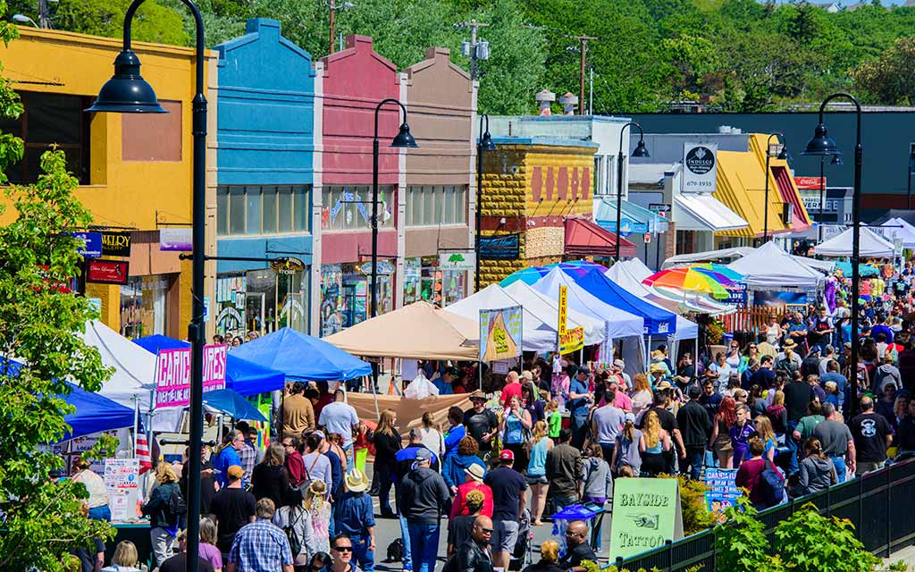 A festival happening in dowtown Oak Harbor. There are many people strolling the road lined with tents and canopies featuring various vendors. In the background are colorful historic buildings in colors of yellow, blue, red and brown.