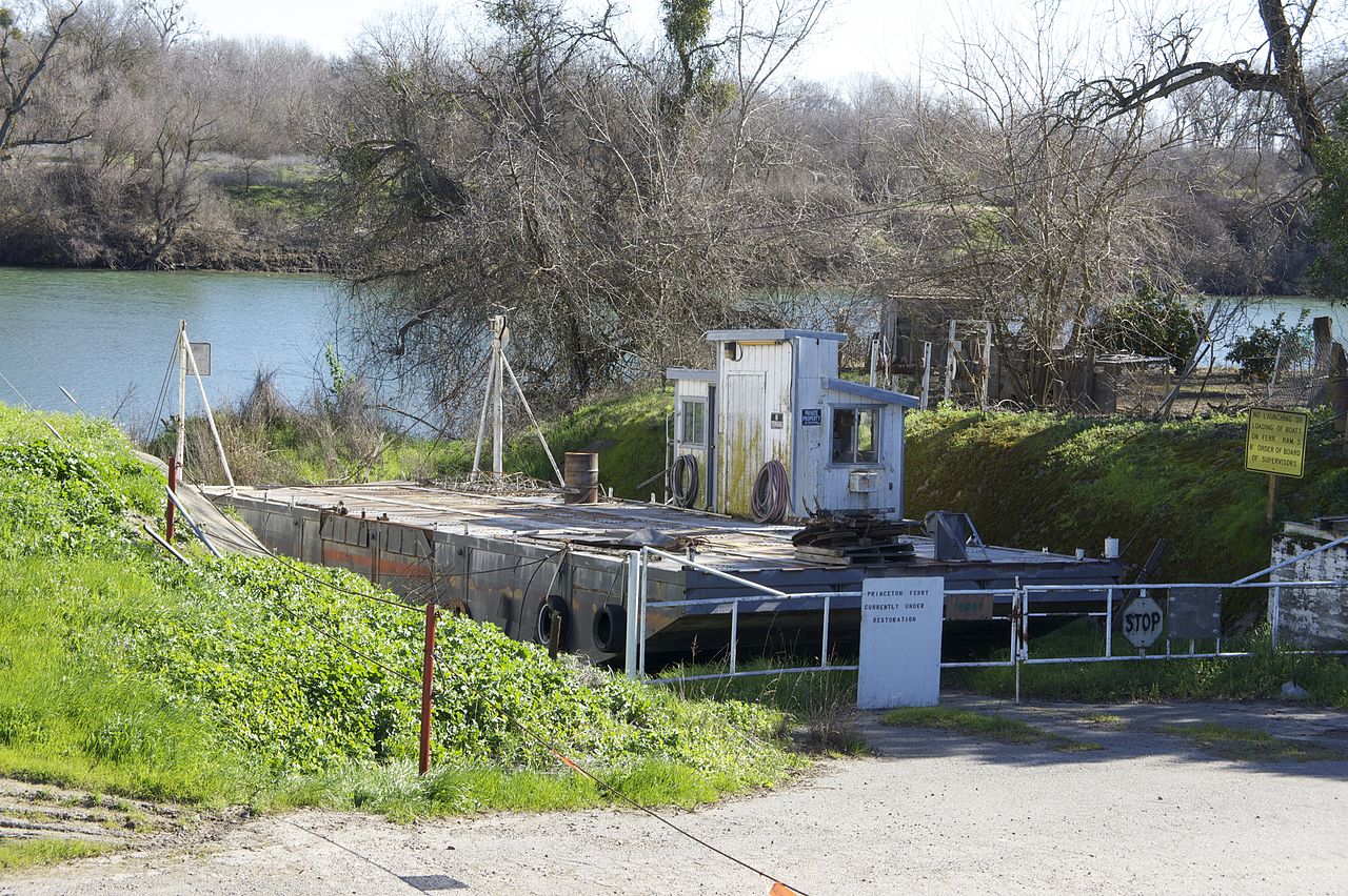 The old Princeton Ferry barge. The abandoned barge is old and rusted and docked on the shore. Overgrown grass and weeds surrounds it. There is a locked, white gate in front of it with a sign that reads Princeton Ferry is currently under renovation. The Sacramento River is in the background.