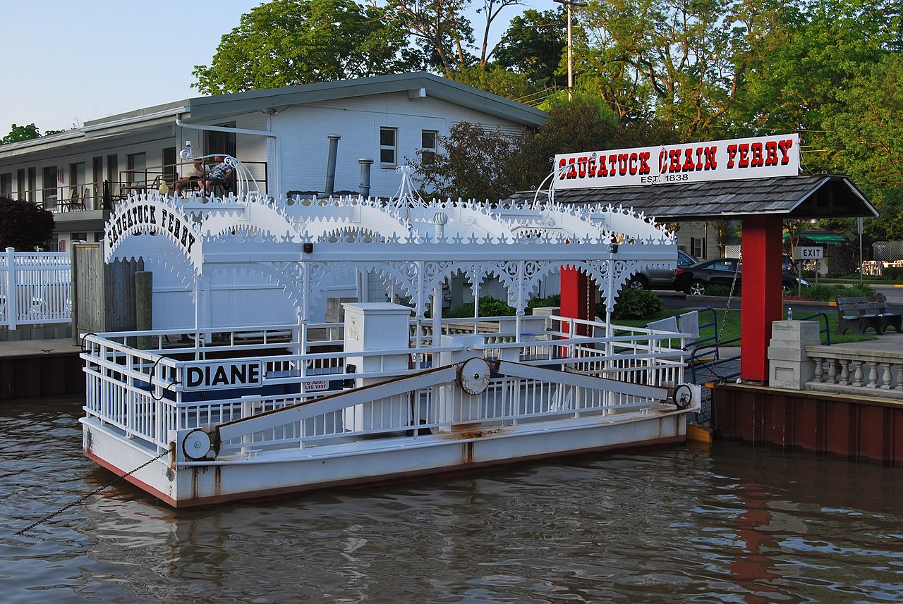 A photo of the Saugatuck Chain Ferry, a type of cable ferry located in Saugatuck, Michigan. The vessel is white and has the name DIANE on the side. The words Saugatuck Ferry adorn the front of the vessel. A gate with a sign on top saying Saugatuck Chain Ferry EST. 1838 is on land behind the vessel. In the background is a two-story motel with balconies on each room. Two people sit in chairs on one of the balconies.