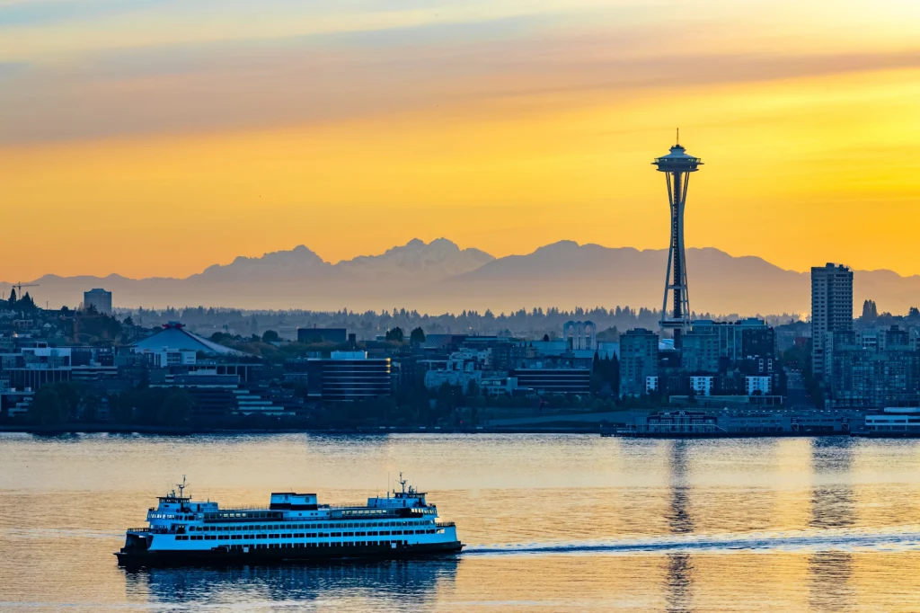 The MV Hyak, a retired Washington State ferry vessel, sails along the water in front of the Seattle skyline during a sunset. The Seattle space needle is visible on the right and the Climate Pledge Arena is visible on the left. The sky is a burning orange color from the sunset.