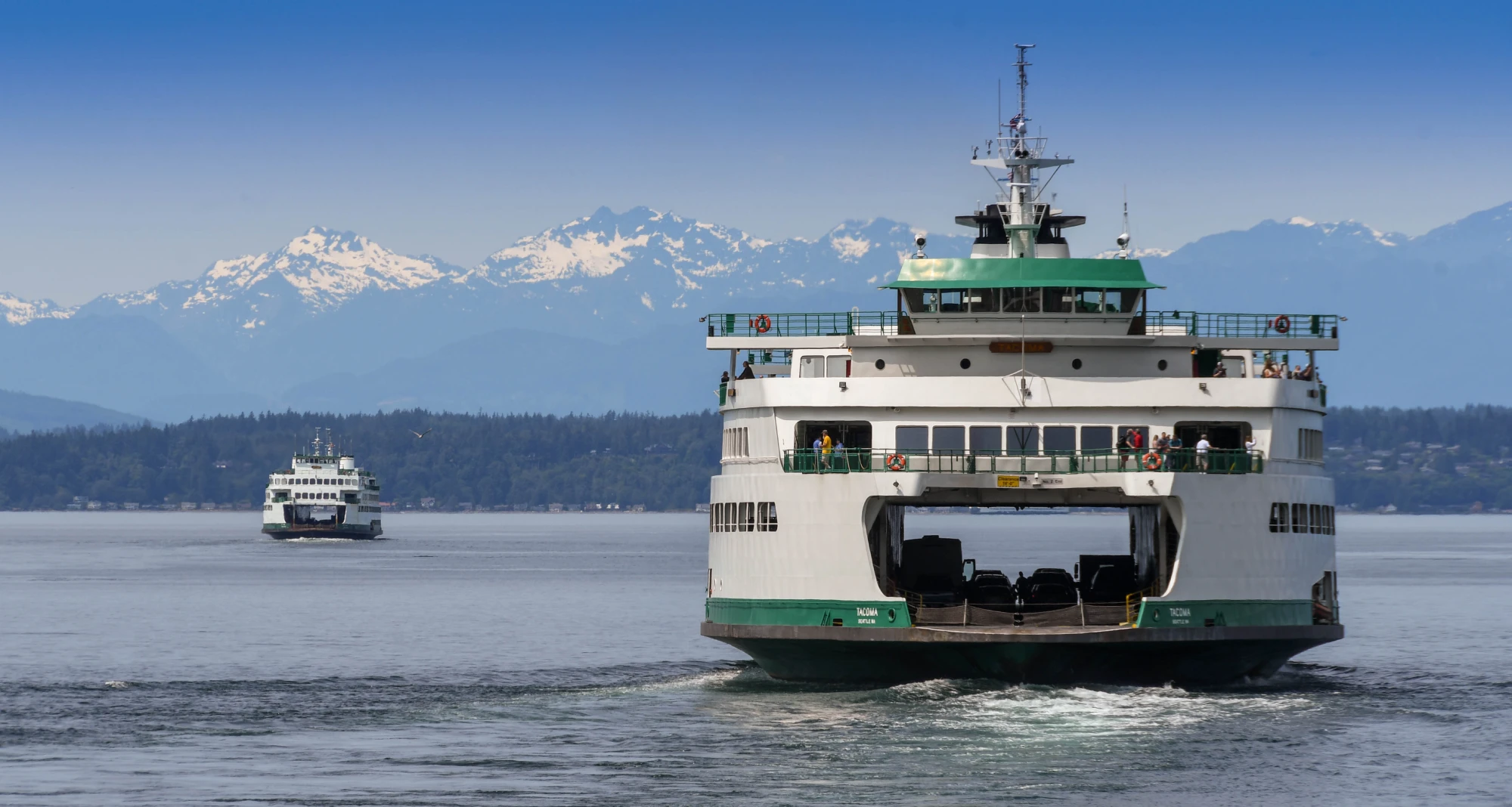 A large passenger and car ferry departs Seattle to cross Puget Sound. In the distance is another passenger and car ferry arriving in Seattle. In the background are the snowcapped Olympic Mountains.