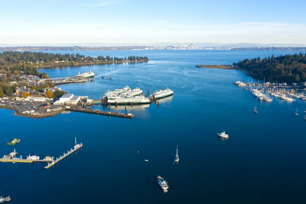 An aerial photograph of the Bainbridge Island ferry terminal. There are four ferry vessels docked at the terminal. A fifth vessel is in the process of docking. There are other boats on the water. Across from a the terminal is a marina with sailboats docked. In the background is Elliott Bay. Beyond Elliott Bay is the Seattle city skyline.