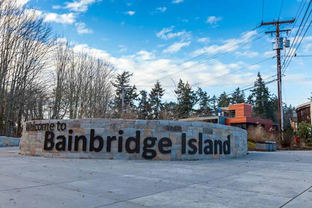 A low angle photograph of a sign that says Welcome to Bainbridge Island in black letters. The sign is constructed of stone and bricks of various colors. On the left side, in the background, are trees with no leaves. On the right side, behind the sign, is a modern looking building. Behind the building are conifer trees.