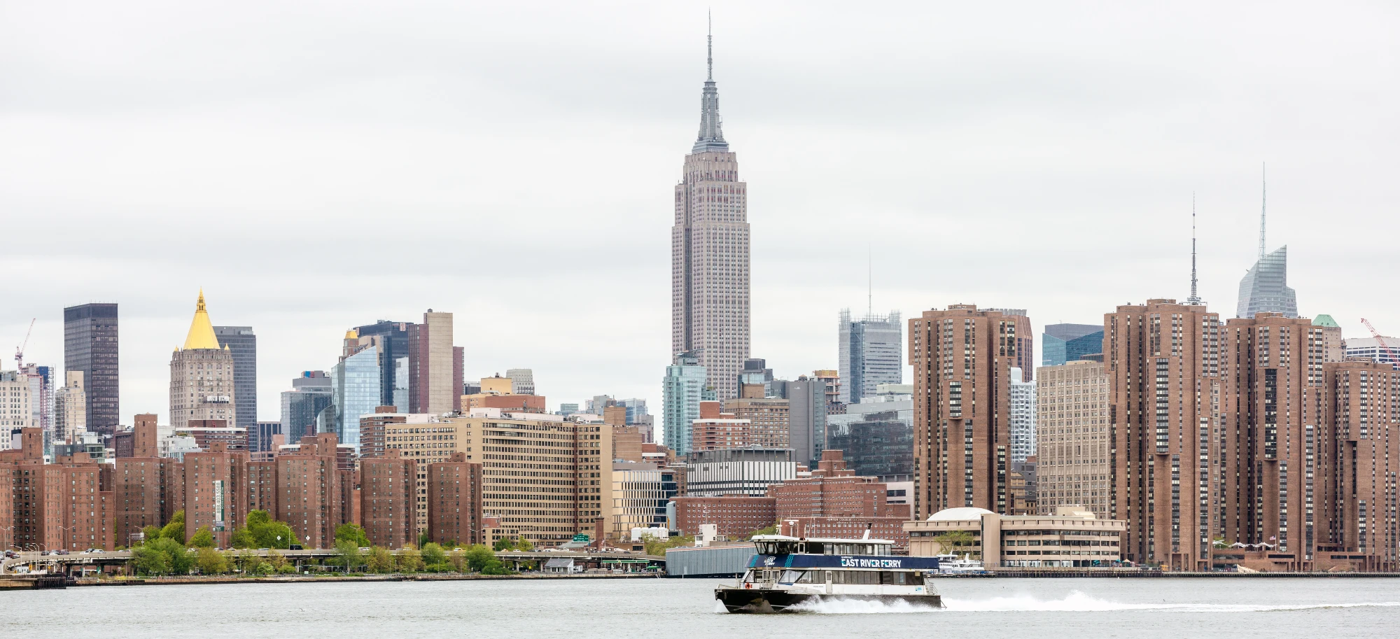 The East River Ferry sails across the East River. In the background is the Manhattan skyline with the Empire State Building in the middle. Off to the right of the skyline is One World Trade Center. It is a gray, overcast day.