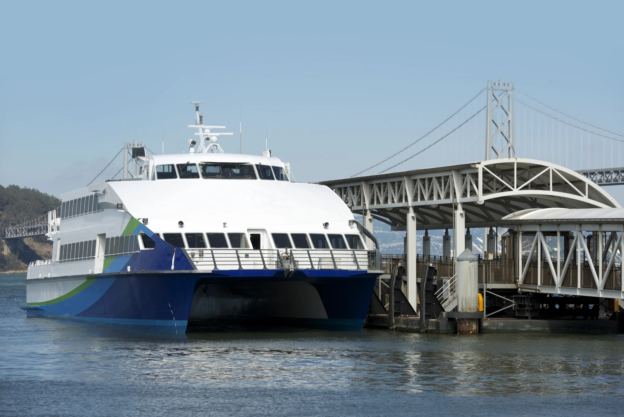 A ferry vessel owned by San Francisco Bay Ferry docks at a pier at the San Francisco Ferry Building. The boat has a primarily white hull with the accent colors of green and blue. It has two passenger decks with windows on each. In the background is the Bay Bridge.