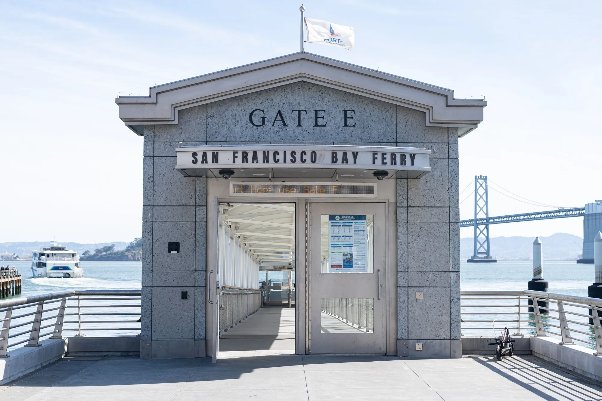 Gate E for San Francisco Bay Ferry at the San Francisco Ferry Building. There are two steel doors on the gate. One of the doors is open. On the left side of the gate a ferry boat departs from the dock. On the right of the gate, in the background, is the Bay Bridge. It is a clear, bright day.