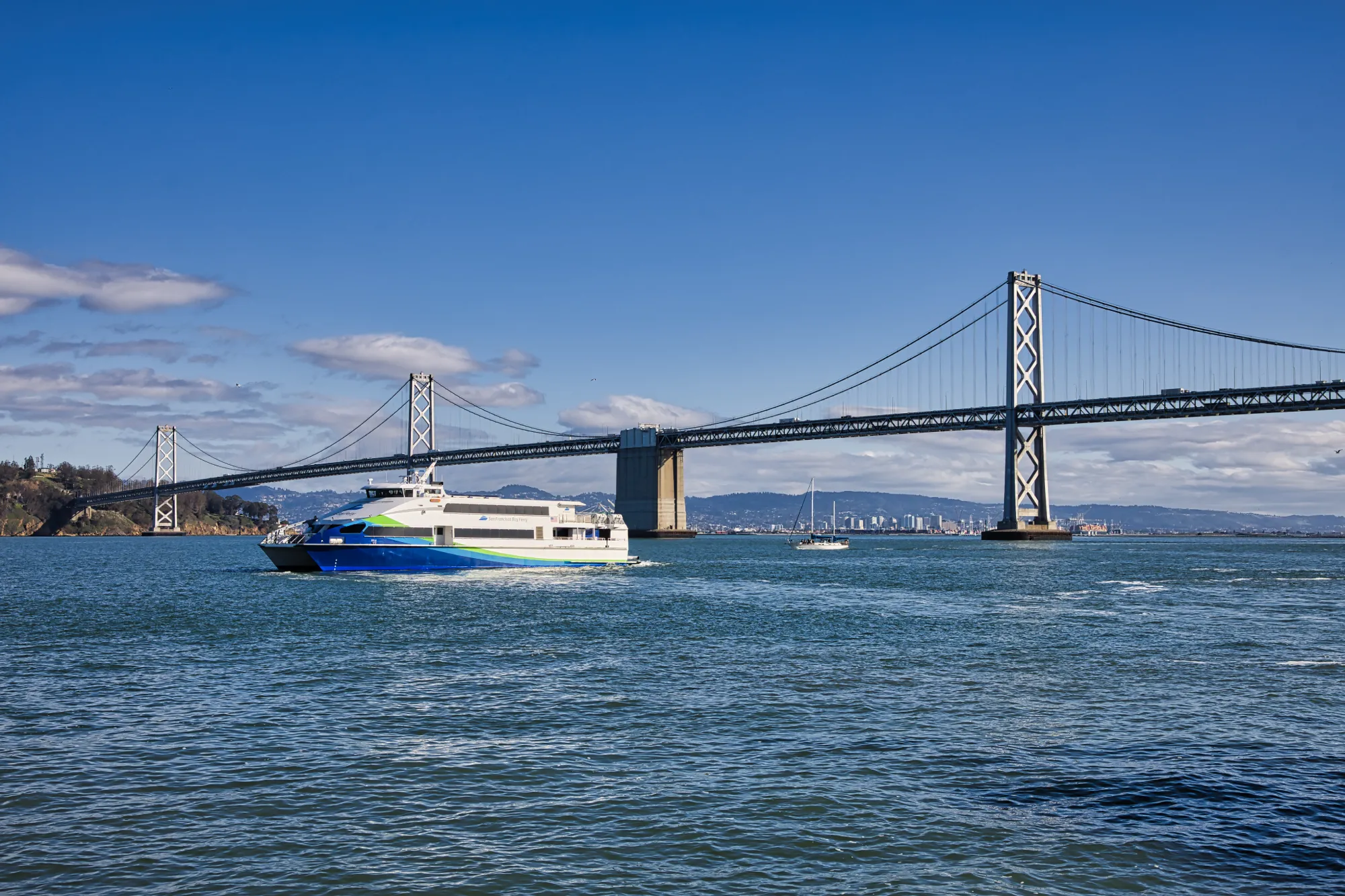 The MV Vela, a ferry vessel operated by San Francisco Bay Ferry, sails across the San Francisco Bay on a sunny, clear day. The Bay Bridge is in the background.